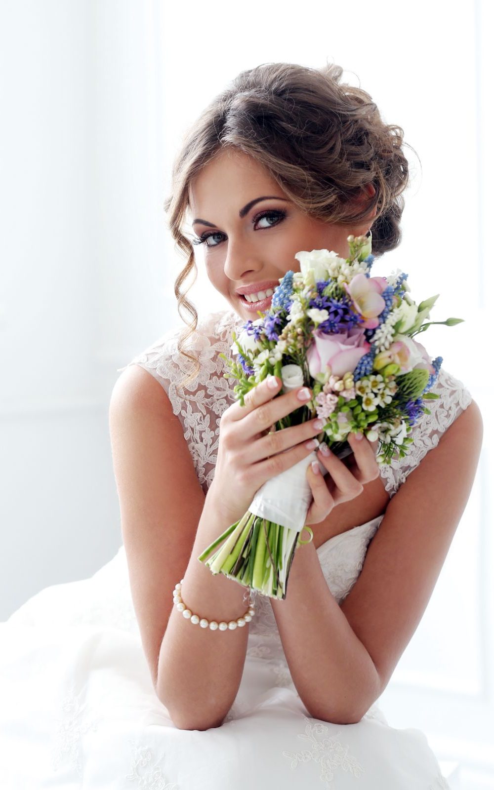 Braut in weißem Spitzenkleid hält lächelnd einen Strauß mit lila und weißen Blumen.